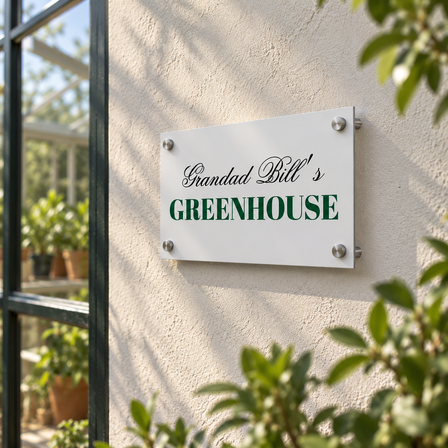 Aluminium garden sign reading Grandad Bill's Greenhouse mounted with wall standoffs on a rendered wall next to a greenhouse