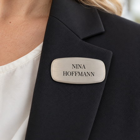 Brushed nickel-plated name badge with pin fastening worn on a dark blazer lapel